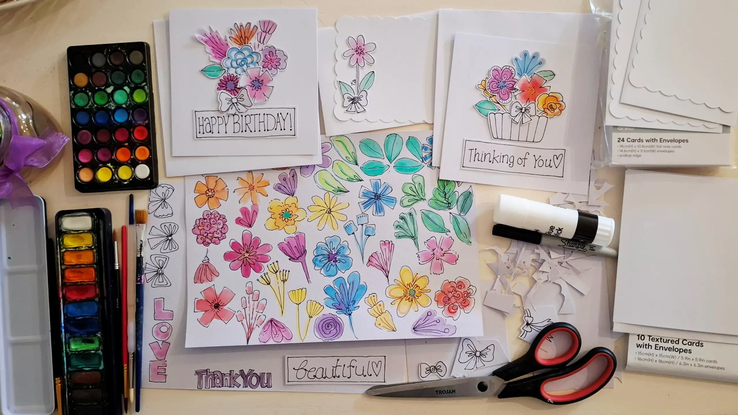 Watercolour card-making workspace with painted floral designs, handmade greeting cards, paint palettes, brushes, scissors, glue and blank cards arranged on a craft table.